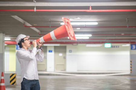 Young Asian engineer man wearing safety helmet yelling though traffic safety cone in parking lot. Construction cone safety conceptの写真素材
