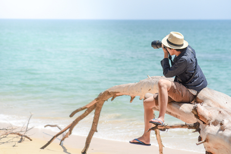 young man photographer with jean shirt and hat sitting on white dead tree near sandy beach of tropical island and taking photo of turquoise sea, summer holiday and vacation travel conceptsの写真素材