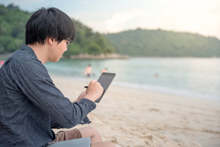 Young Asian man using tablet on tropical beach, digital nomad or freelance lifestyle conceptsの写真素材