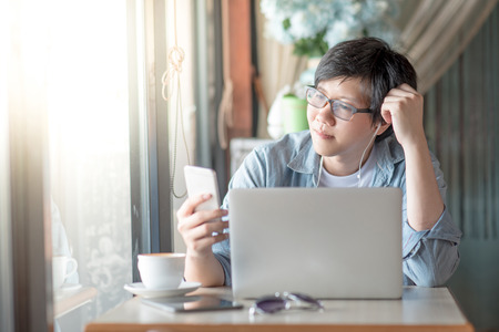 Young Asian man listening to music on moblie application. Casual professional entrepreneur using smartphone in cafe or coffee shop. modern lifestyle and digital age conceptsの写真素材