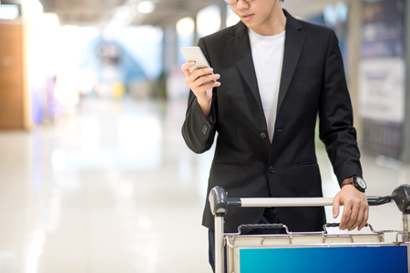 Young asian man with his suitcase luggage using smartphone while waiting for airline flight in the international airport terminal, business travel and online check in conceptsの写真素材