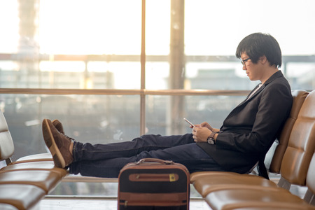 Young Asian well-dressed businessman using smartphone sitting on bench near his suitcase luggage while waiting for connecting flight in airport terminal. man in business travel conceptの写真素材