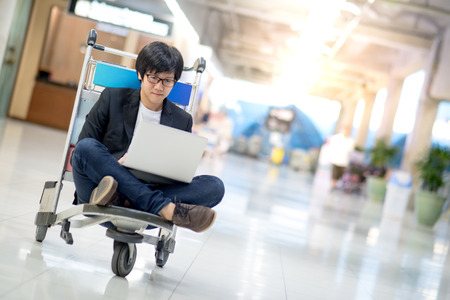 Young asian man working on airport trolley with his laptop computer during waiting for a connecting flight, freelance lifestyle and digital nomad conceptsの写真素材