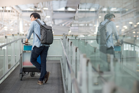 Young Asian man walking with airport trolley and his suitcase luggage in the international airport terminal, arrival from travel abroadの写真素材