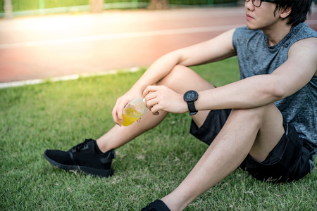 Young Asian man runner sitting on green grass drinking energy drink after jogging in the park. healthy lifestyle with urban running conceptの写真素材