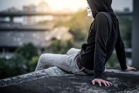 Mystery man in white mask wearing hoody sitting on rooftop of abandoned building feeling lonely, self destruction suicidal addiction or major depressive disorder conceptsの写真素材