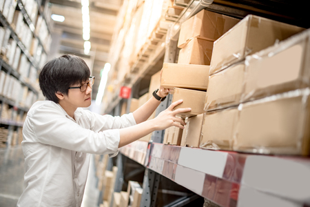 Young Asian man picking paper box from shelf in warehouse, shopping warehousing or packing products conceptsの写真素材