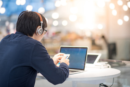 Young Asian business man listening to music by headphones and smartphone while working with laptop computer in co working space. freelance or digital nomad lifestyle in urban workspace conceptの写真素材