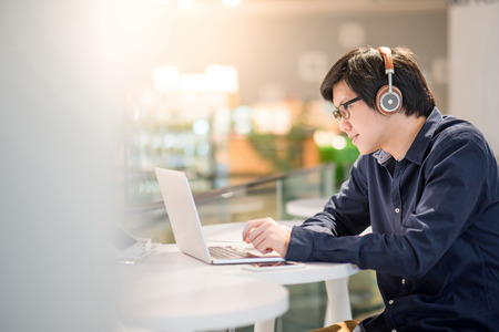 Young Asian business man listening to music by headphones while working with laptop computer in co working space. freelance or digital nomad lifestyle in urban workspace conceptsの写真素材