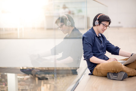Reflection of young Asian business man enjoy listening to music by headphones while using laptop computer in co working space. freelance or digital nomad lifestyle in urban workspace conceptsの写真素材