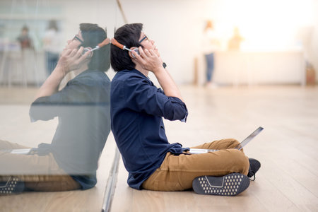 Reflection of young Asian business man enjoy listening to music by headphones while using laptop computer in co working space. freelance or digital nomad lifestyle in urban workspace conceptsの写真素材