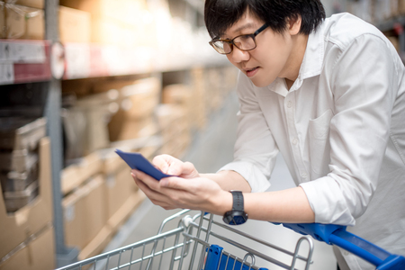 Young Asian man standing with cart checking the shopping list in warehouse wholesale, shopping warehousing conceptの写真素材