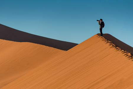 Young male traveler and photographer standing on the top of sand dune photographing sunrise or sunset in desert of Namibia, Africa. Travel photography conceptの写真素材