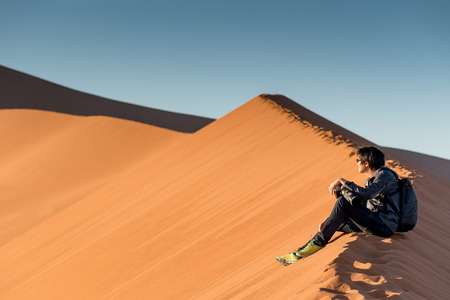 Young male traveler and photographer sitting on the top of sand dune looking at sunrise or sunset in desert of Namibia, Africa. Travel photography conceptの写真素材