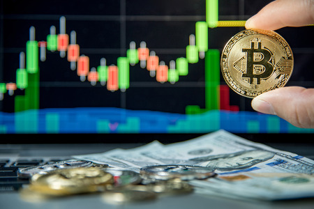 Male hand showing gold bitcoin. Dollar bank note and coins on laptop keyboard with basic candlestick green red graph price in background. Worldwide cryptocurrency and digital payment conceptsの写真素材