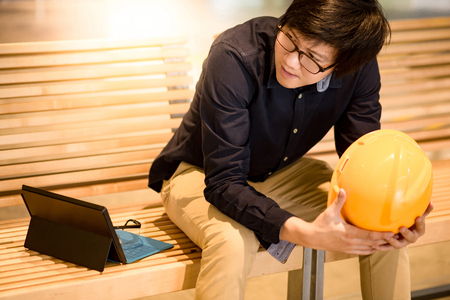 Young Asian engineer man holding personal yellow protective safety helmet while sitting with digital tablet and safety glasses on wooden bench. Male architect, Construction and Engineering conceptsの写真素材