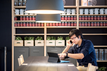 Young Asian businessman using smartphone for business talk pointing on his digital tablet in co working space. freelance lifestyle or digital nomad conceptsの写真素材