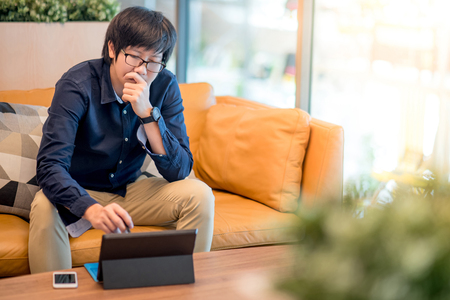 Young Asian businessman feeling stressed and frustrated while working with digital tablet on sofa in living room. Business problem conceptの写真素材