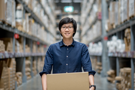 Young Asian man carry cardboard box between row of shelves in warehouse, shopping warehousing or working pick and packing conceptsの写真素材