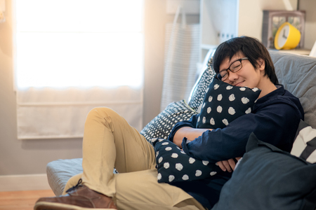 Young Asian man with glasses hugging cushion sleeping on sofa in living room. Lifestyle in living space conceptの写真素材