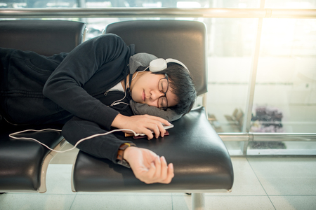 Young Asian man with eyeglasses and headphones sleeping on bench while waiting for connecting flight in the international airport terminal, travel lifestyleの写真素材