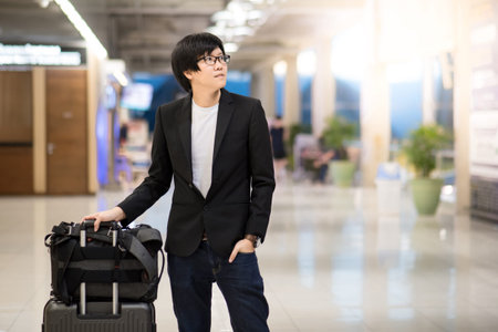 Young asian businessman with his suitcase luggage and backpack waiting for airline flight in the international airport terminal, man in business travel conceptの写真素材