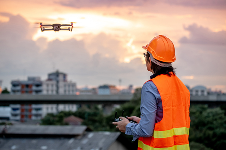 Young Asian engineer flying drone over construction site during sunset. Using unmanned aerial vehicle (UAV) for land and building site survey in civil engineering project.の写真素材