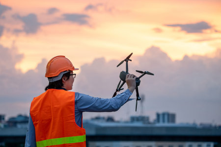 Young Asian engineer holding drone at construction site during sunset. Using unmanned aerial vehicle...
