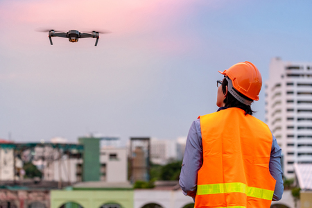 Young Asian engineer flying drone over construction site during sunset. Using unmanned aerial vehicle (UAV) for land and building site survey in civil engineering project.の写真素材