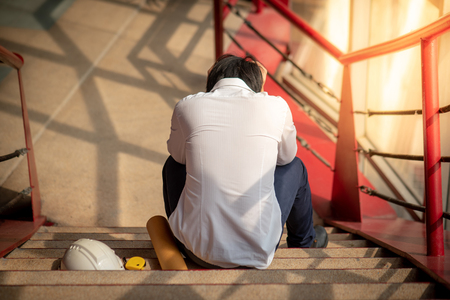Engineer or Architect feeling stressed, worried and tired with his job while sitting on stair with protective safety helmet at construction site.  Problem and failure with construction project conceptの写真素材