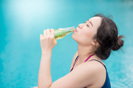 Young Asian attractive woman drinking sport drink water after do yoga at swimming pool, outdoor...
