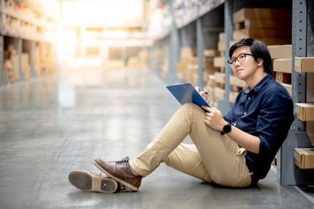 Young Asian man doing stocktaking of product in cardboard box on shelves in warehouse by using clipboard and pen. physical inventory count conceptの写真素材