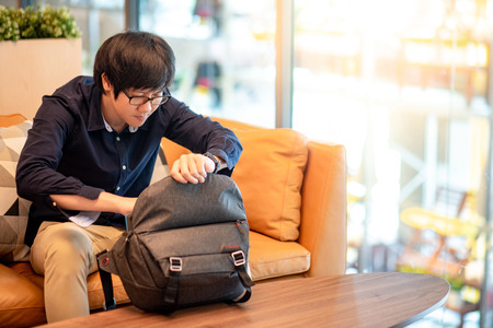 Young Asian man putting digital tablet into his bag sitting on sofa in living room. Urban lifestyle conceptの写真素材