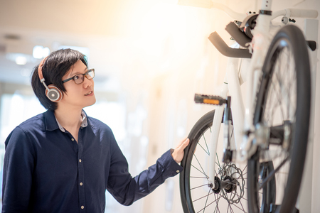 Young Asian man with headphones and glasses looking for bicycle at bike shop in department store, urban lifestyle in shopping mall conceptの写真素材