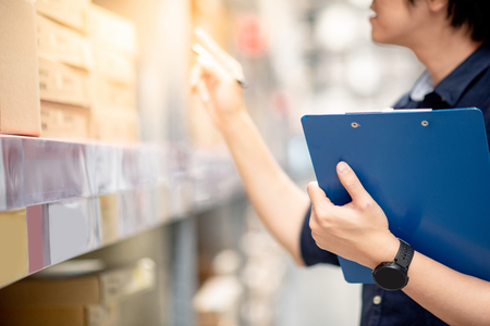 Young Asian man doing stocktaking of product in cardboard box on shelves in warehouse by using clipboard and pen. physical inventory count conceptの写真素材