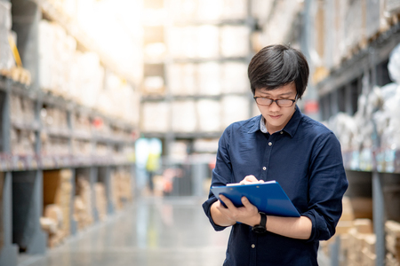 Young Asian man doing stocktaking of product in cardboard box on shelves in warehouse by using clipboard and pen. physical inventory count conceptの写真素材