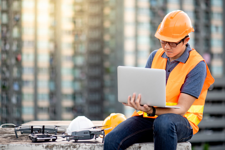 Young Asian man working with drone laptop and smartphone at construction site. Using unmanned aerial vehicle (UAV) for land and building site survey in civil engineering project.の写真素材