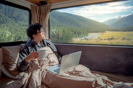 Young Asian man drinking hot coffee working with laptop computer on the bed in camper van with mountain scenic view through the window, digital nomad on road trip conceptの写真素材