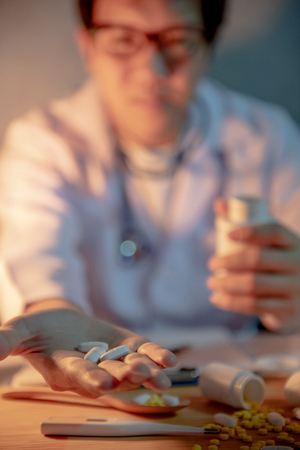 Male Asian doctor showing pills on his hand. Doctor working with medicine paperwork on the desk in clinic or hospital. Medical treatment and health care concepts.の写真素材
