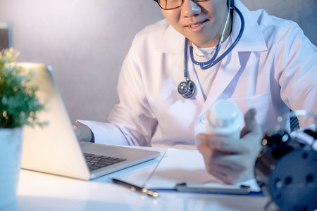 Male Asian doctor looking at pill bottle on his hand working with laptop computer and medicine paperwork clipboard on desk in clinic or hospital. Medical treatment and health care concepts.の写真素材