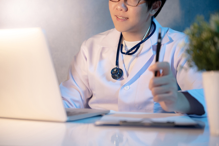 Male Asian doctor working with laptop computer and medicine paperwork clipboard on the desk in hospital clinic. Medical treatment and health care consultation concepts.の写真素材