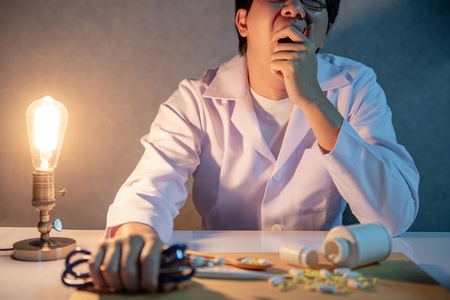 Overworked doctor yawning while sitting with stethoscope and medicine in hospital clinic. Male practitioner feeling stressed and tried during hard working time in medical center.の写真素材