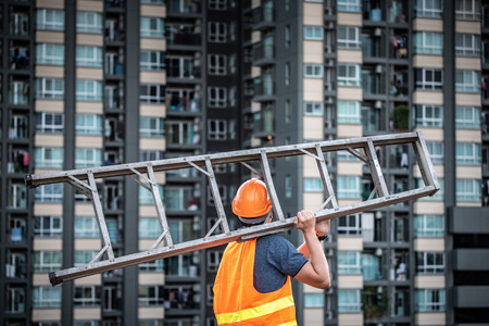 Young Asian maintenance worker with orange safety helmet and vest carrying aluminium step ladder at construction site. Civil engineering, Architecture builder and building service conceptsの写真素材