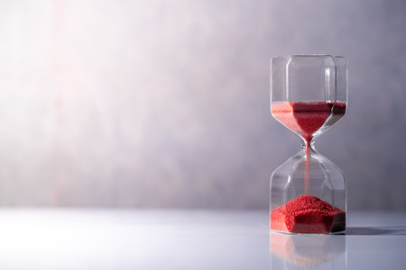 Red sand running through the shape of modern hourglass on white table.Time passing and running out of time. Urgency countdown timer for business deadline concept with copy spaceの写真素材