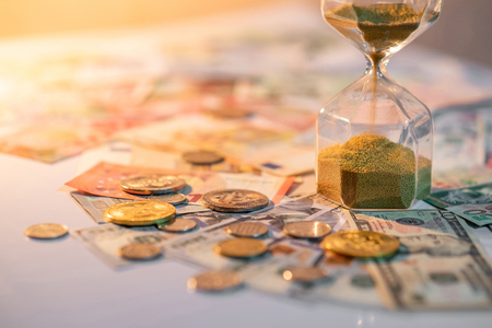 Sand running through the shape of hourglass on table with banknotes and coins of international currency. Time investment and retirement saving. Urgency countdown timer for business deadline conceptの写真素材