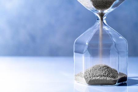 Sliver sand running through the shape of modern hourglass on white table.Time passing and running out of time. Urgency countdown timer for business deadline conceptの写真素材