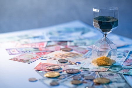 Sand running through the shape of hourglass on table with banknotes and coins of international currency. Time investment and retirement saving. Urgency countdown timer for business deadline conceptの写真素材