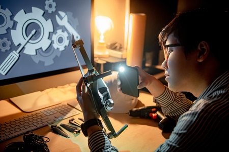 Asian technical engineer using flashlight from smartphone for checking drone body. Male technician fixing or maintenance drone. Unmanned aerial vehicle (UAV) photography conceptの写真素材