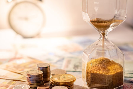 Sand running through the shape of hourglass on table with banknotes and coins of international...