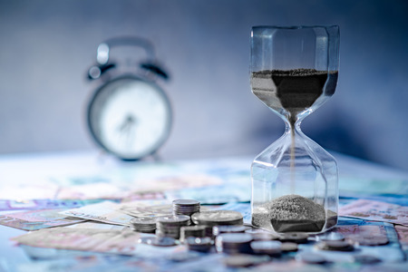 Sand running through the shape of hourglass on table with banknotes and coins of international currency. Time investment and retirement saving. Urgency countdown timer for business deadline conceptの写真素材